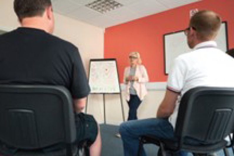 Two people sat in a room facing a third person stood up talking to the room.
