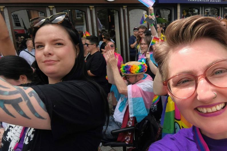 Bonnie has short cropped hair and is taking a selfie with a women who has black hair and is wearing all black