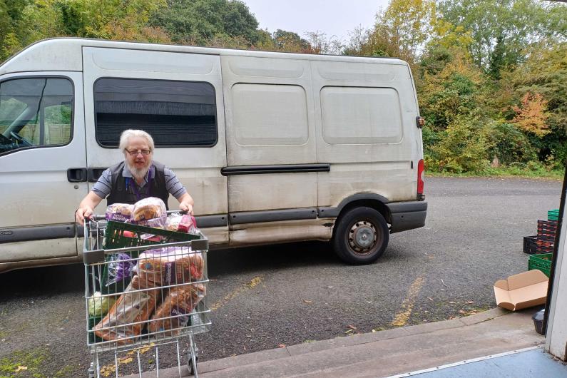 A man pushing a shopping trolley full of food into a warehouse. A white van is parked behind him