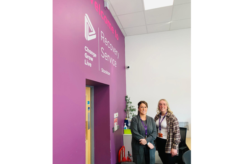 Two women stood next to a pink wall with a big Welcome to Stockton Recovery Service sign on the wall