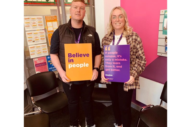 Two people stood in front of a pink wall holding up signs that read 'Believe in People'